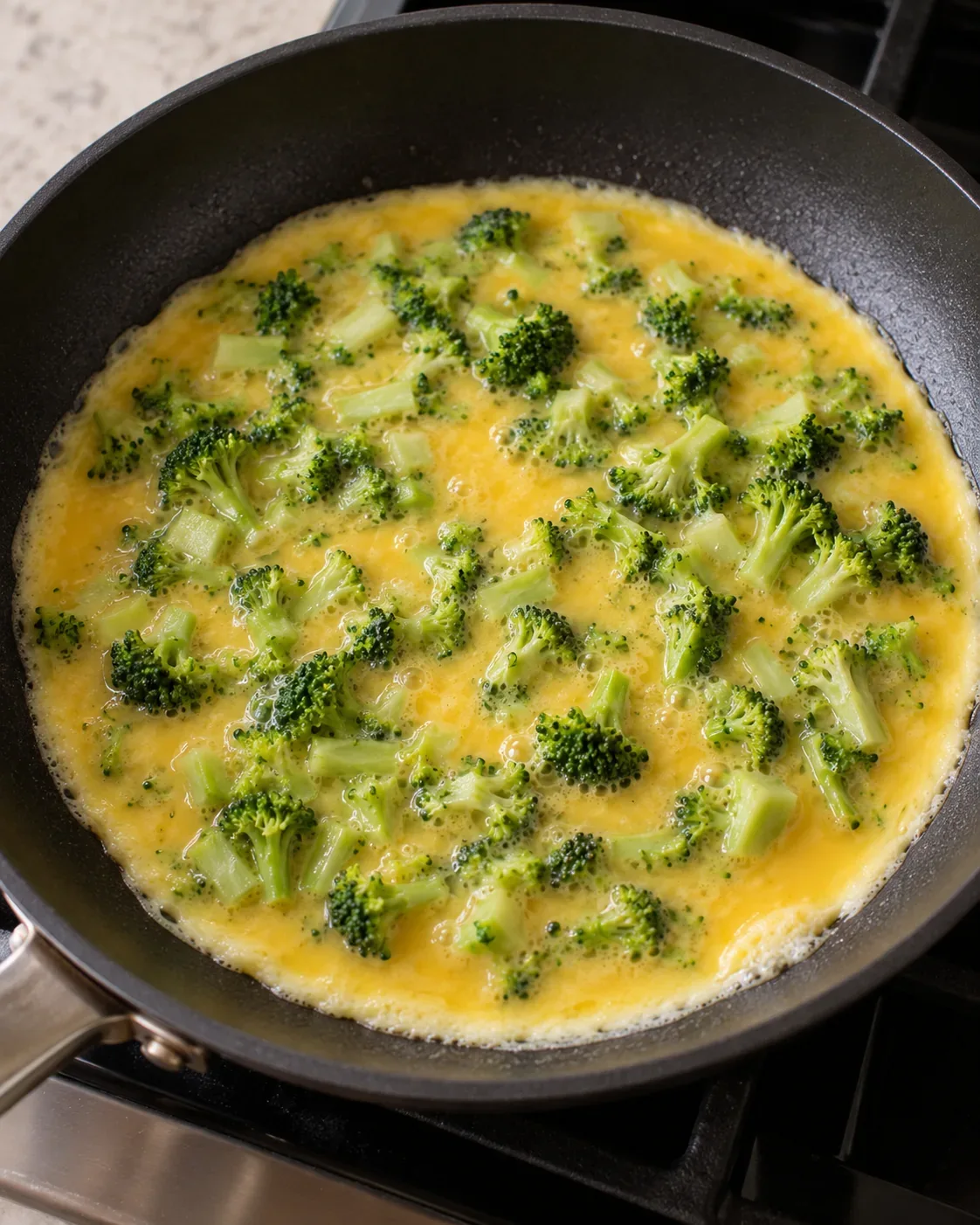 Broccoli egg tortilla fold in progress, with chopped broccoli added to whisked eggs as they begin to set in a skillet.