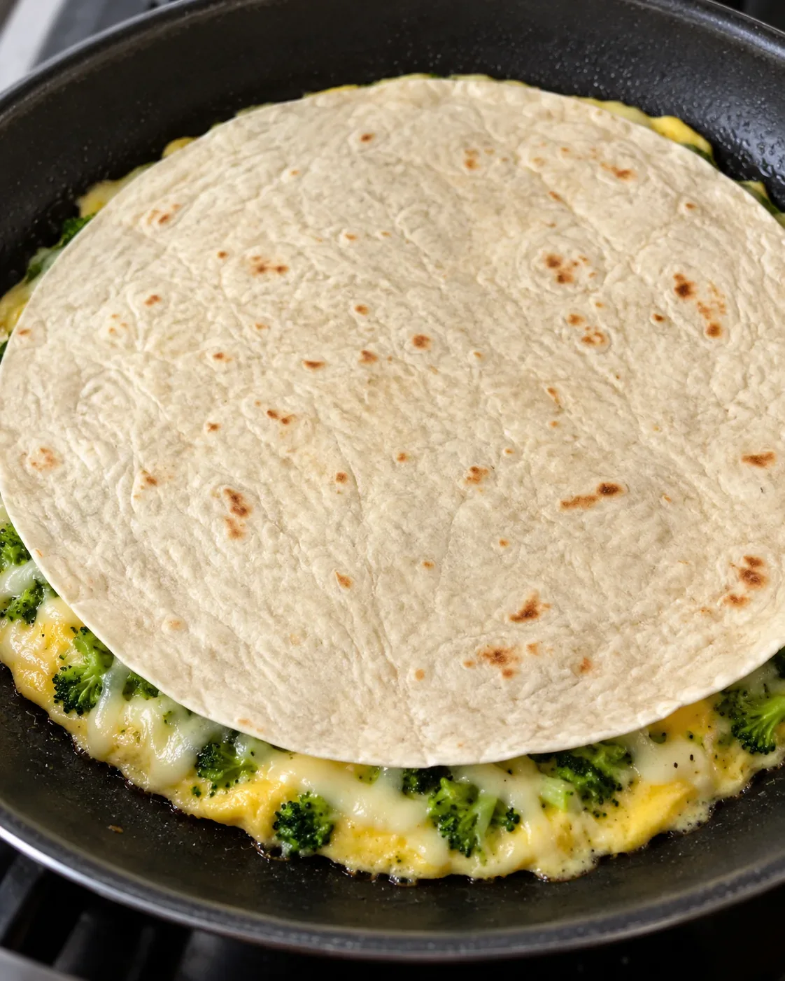 Broccoli egg tortilla fold just before flipping, with a flour tortilla resting over the egg, chopped broccoli, and white mozzarella in the skillet.