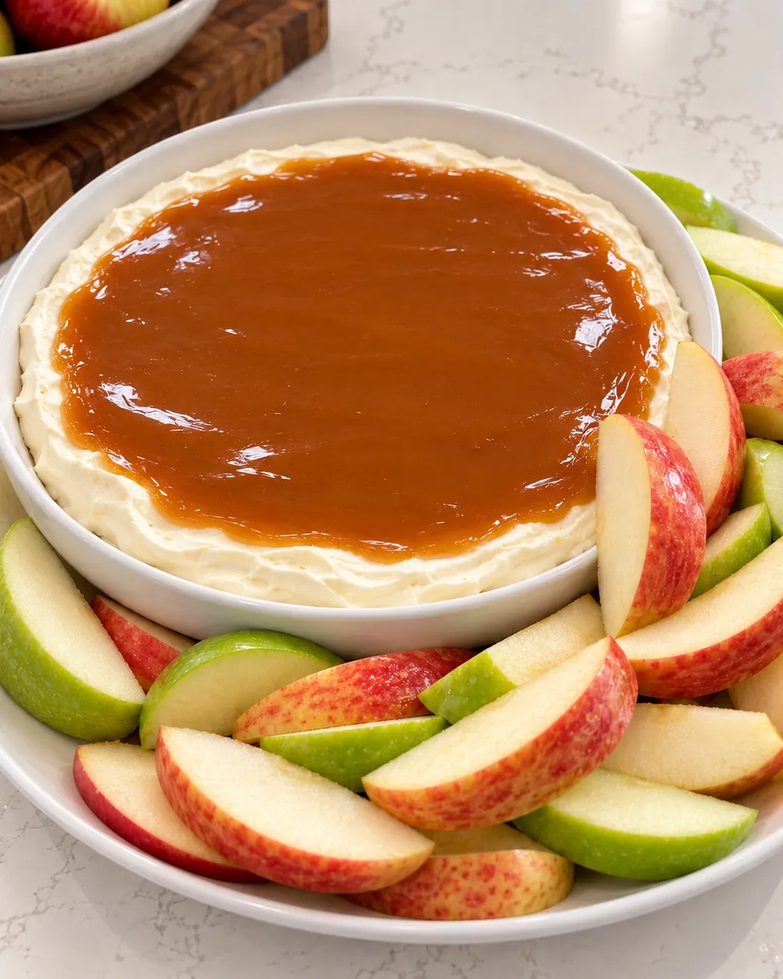 Cream cheese caramel apple dip in a shallow white dish with red and green apple slices.