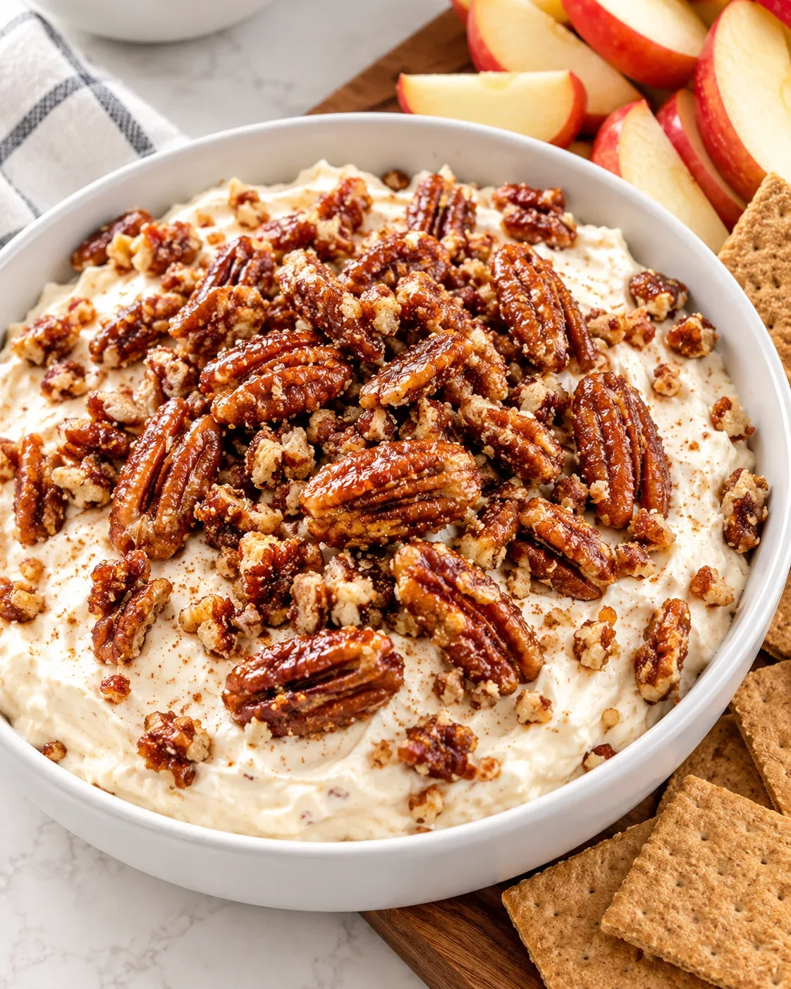 White bowl of creamy dip topped with golden candied pecans, with apple slices and graham crackers nearby.