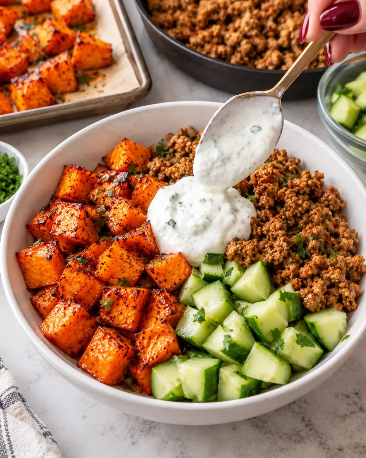 Lemon yogurt being spooned over ground turkey, roasted sweet potatoes, and cucumber in a white bowl.