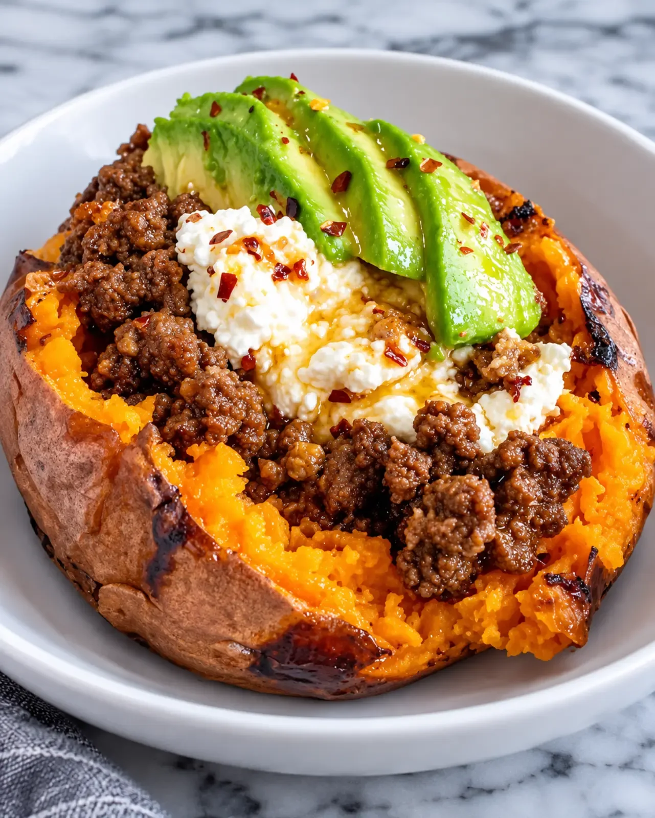 A baked sweet potato filled with seasoned ground beef, cottage cheese, avocado slices, and hot honey.